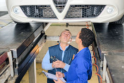 Two mechanics in a vehicle pit beneath a car being serviced.