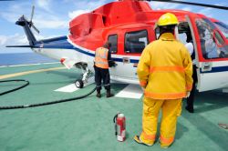 Two workers near helicopter; one fueling the other nearby with a fire extinguisher.  Another
                                        man in a yellow coat and pants stands in the foreground.