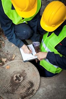 Two workers writing on a clipboard both have helmets and safety vests. Standing near a round sewer cover.