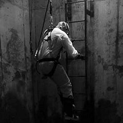 Confined space entrant climbing down a ladder and wearing a harness, hazmat suit, and respirator and retrieval line.