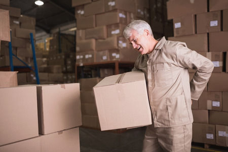 Older worker lifting a box in a warehouse and holding his back because he hurt it.