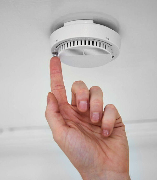 A person's finger touching a smoke detector test button mounted to the ceiling