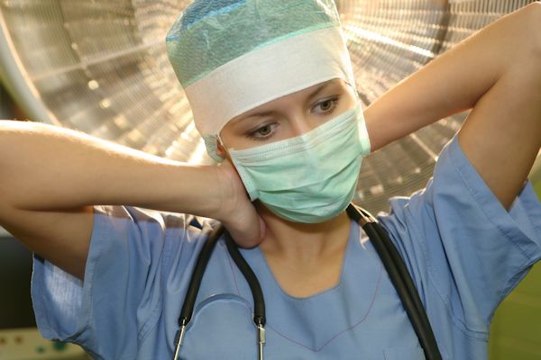 Tired doctor wearing scrubs, face mask, and stretching back and neck.