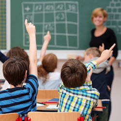 Teacher stands in front of class with a chalkboard in the background. Students in the class have their hands raised.