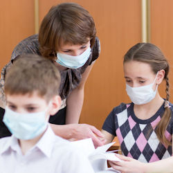 Two students sitting at desks. Teacher is leaning over student in the back. The teacher and students are wearing masks that cover their nose and mouth