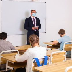 Students sitting at desk facing the teacher who is standing at the front of the room. Everyone is wearing masks. The teacher is in front of a white board.