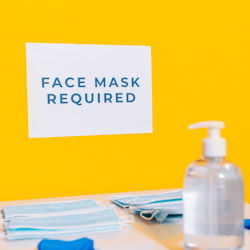 Sign on wall reading 'Face Mask Required'. The sign is white with blue text. The wall is yellow. There is a bottle of hand sanitizer and face masks on a table infront of the sign.