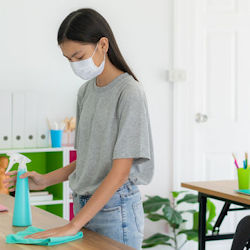 A woman wearing a mask cleans a wooden table using a spray bottle and a rag.