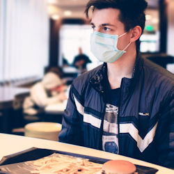 Student, wearing a mask, sits at a lunch table. There is lunch tray with a burger and fries sitting on the table in front of him.