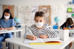 Girl wearing a striped shirt and a face mask sits at desk working in a book. There are two other girls behind her sitting at desks that are slightly out of focus.