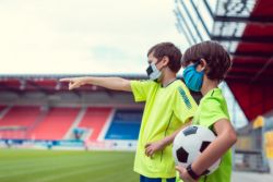 Two children, wearing bright green jerseys, stand on a field in a stadium. Both are wearing face masks. One of the children is holding a soccer ball.