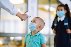 A small child, wearing a turquoise shirt, has his temperature taken. There is another child standing behind him waiting her turn.