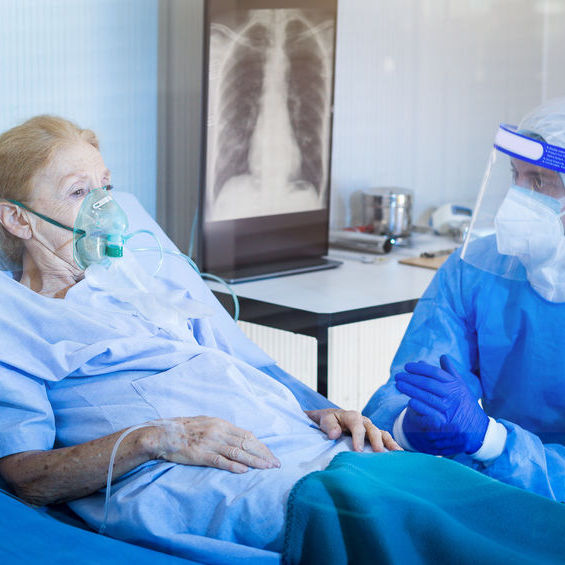 man wearing scrubs, face mask, and a face shield sits at the bedside of a female patient. The female patient is wearing a hospital gown and oxygen mask. An x-ray is posted on the wall