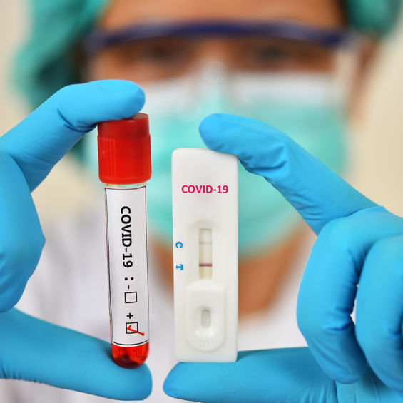 A tube of blood and a COVID-19 test strip being held by two gloved hands.