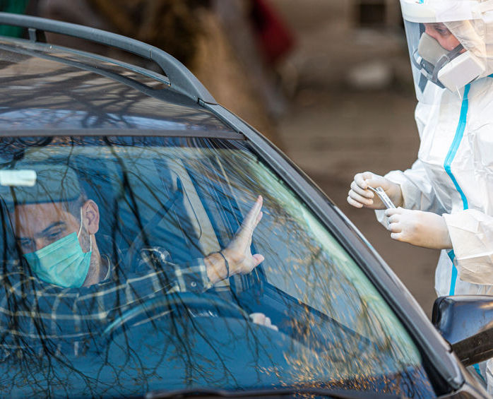 A man sits in a car wearing a face mask. He is holding his hand up in a no/stop posiiton. Nurse in full PPE standing outside car