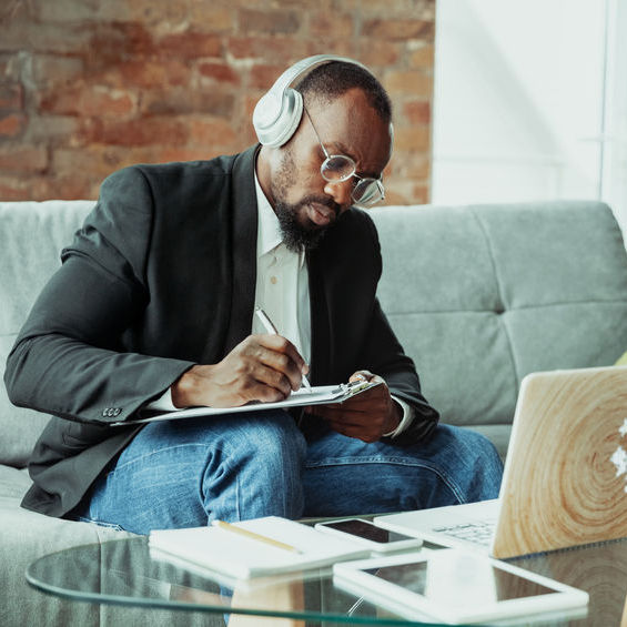 A man sitting on a couch wearing headphones, fills out paperwork on a clipboard.