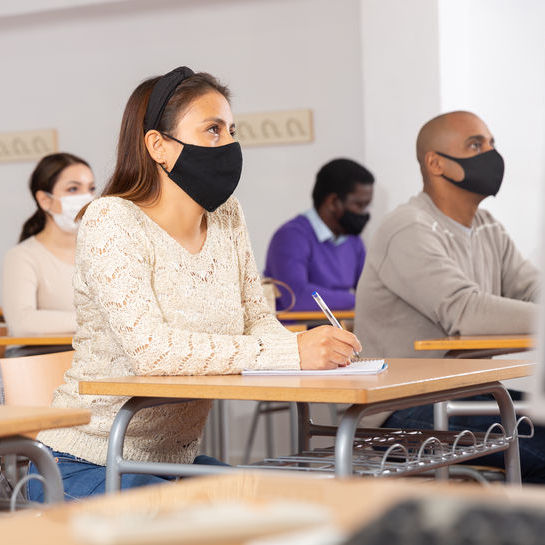 Four students, wearing face masks, sitting at desks taking notes in a classroom.