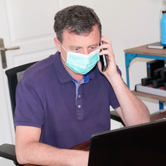 Man wearing a facemask sitting at a desk in front of a computer talking on the phone.