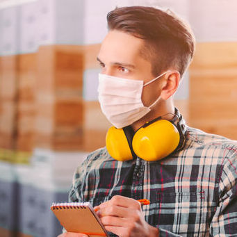 A man wearing a face mask is taking notes on a notepad. He has hearing protection resting around his neck.