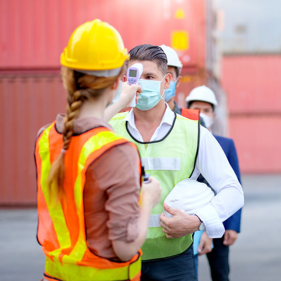 A woman in a hard hat and safety vest wearing a
                                        facemask uses a forehead thermometer to take a mans temperature. The man is wearing a safety vest and a face mask.