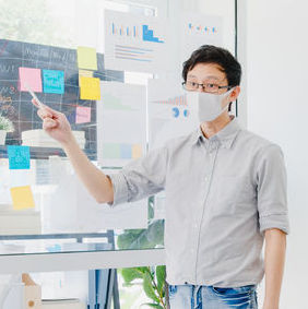 A man wearing a face mask points to a board with
                                        sticky notes and papers taped to it.