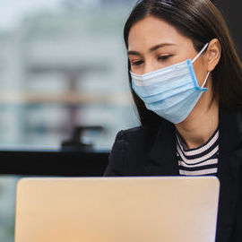 A woman wearing a facemask with a laptop computer screen opened in front of her.