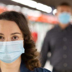 Woman wearing a face mask. There is a man in the background wearing a face mask, but he is not in focus.