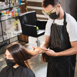 A stylist is looking at a womans hair. The woman has a gown
                                        and face mask on. The stylist is wearing an apron, face mask, and face shield.