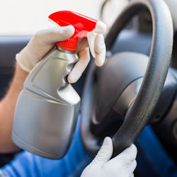 A person wearing latex gloves uses a spray bottle to clean a steering wheel in a car.