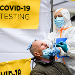 A man gets a COVID-19 test. The person administering
                                        the test are in full PPE. The man getting the test done has a face mask pulled down around his chin.