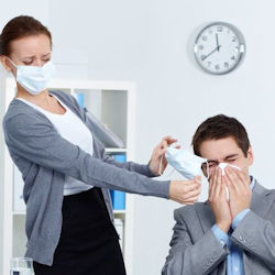 A woman wearing a face mask tries to hand a face
                                        mask to a man. The man is sitting in a chair blowing his nose.