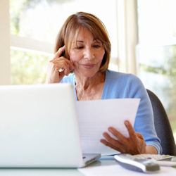 A woman sits at a desk looking at a piece of paper. There is a laptop in front of her.