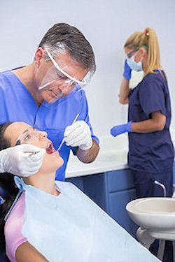 Dentist wearing gloves and a face shield inspecting a patients teeth. Nursing assistant in the background in PPE