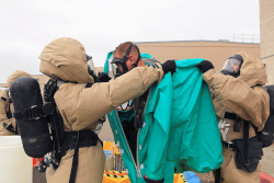 Two workers in Level B protective suits removing a Level A protective suit from a worker that was recently decontaminated