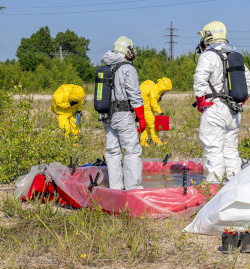 Two people in gray protective suits with air supply stand in a decontamination area. There are two people in yellow suits in the background removing hazardous substances.