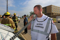 A man speaks into a radio. He is wearing a white Incident Commander vest. There are emergency response workers behind him.