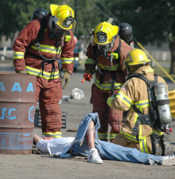 Firefighters in proctective suits, helmets, and respirators attend to a victim laying on the ground.