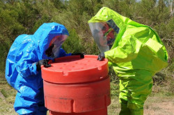 A person in a blue hazmat suit and a person in a bright green hazmat suit inspect a container of hazardous waste