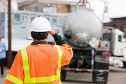 A man in a white hard hat and an orange safety vest has his back to his camera. He is observing a truck.