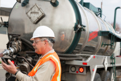 A man, wearing an orange safety vest and a white hard hat, stands behind a truck