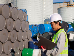 A person, wearing a green safety vest and a white hard hat, carries a clipboard while inspecting stacked barrels. 