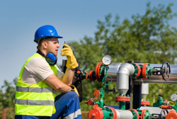 A man, wearing a blue hard hat and a green safety vest talks into a 2-way radio. He has blue ear protection around his neck and is standing next to a number of pipes.  