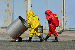 A person in a red hazmat suit and a person in a yellow hazmat suit push an empty container on a hand truck.