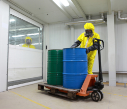 Man in a yellow protective suit and respirator moves two barrels using a pallet jack.