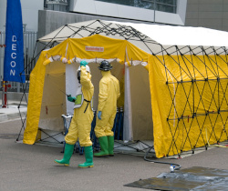 Two workers in yellow suits with hard hats stand outside a decontamination shower