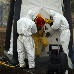 Two men in white hazmat suits help a man, wearing a red hardhat and yellow suit, out of a decontamination tent.