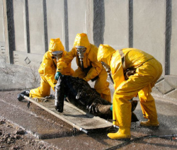 Three men in yellow hazmat suits move a dummy onto a stretcher.