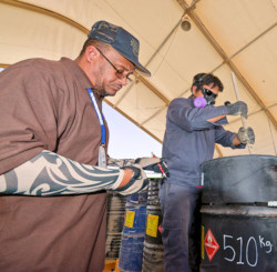 Man takes notes while another man, wearing a respirator, uses a stick to check for sediments inside a drum.