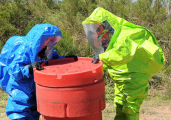 One person in a blue hazmat suit and another person in a green hazmat suit lift the lid off a container.