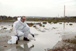 Man in white protective suit takes a water sample from an outdoor pool of water.